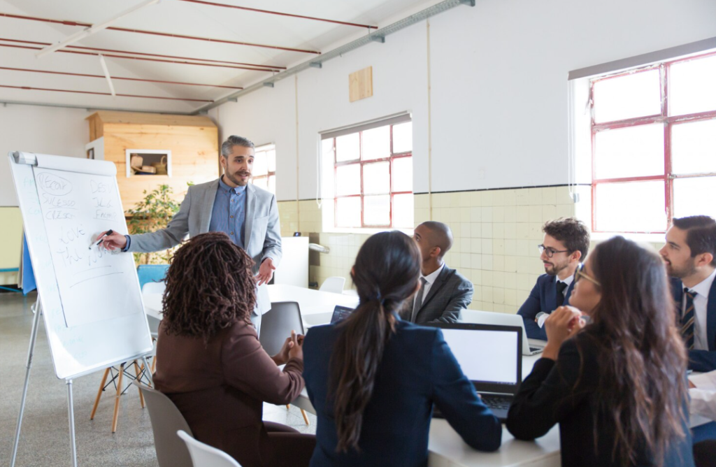 Employees sitting around a table with their laptops open watching a man present ideas on a white board
